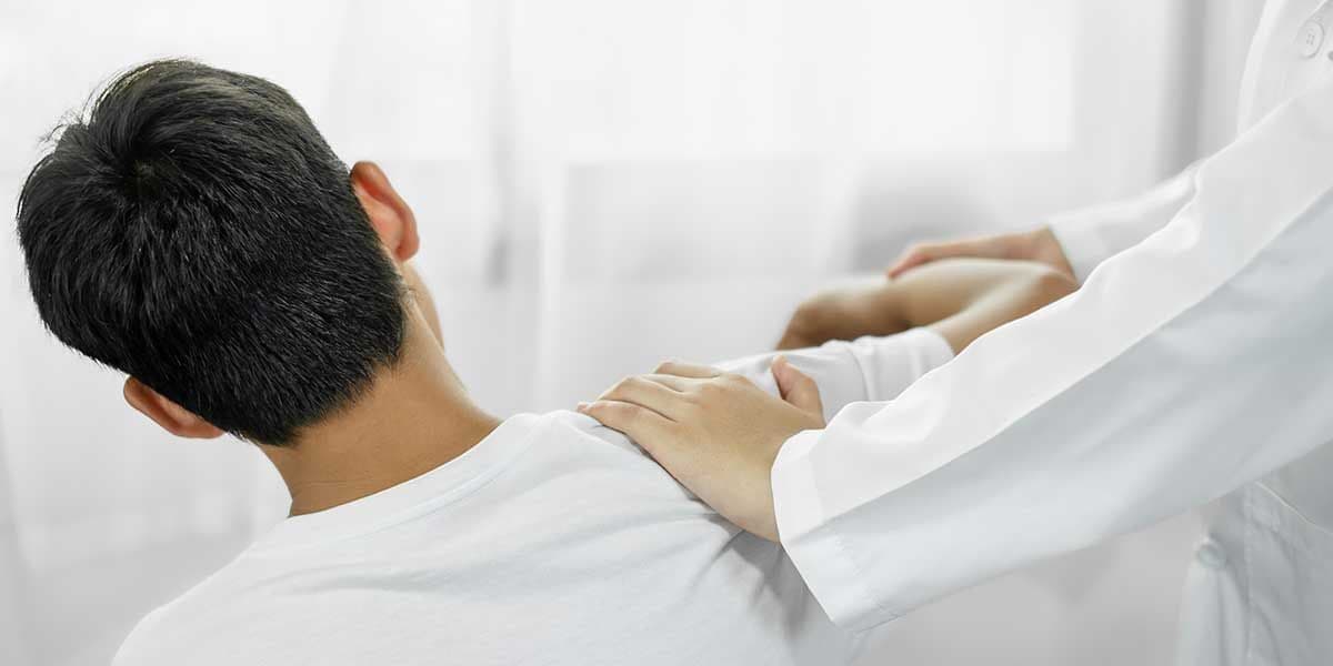 A medical professional in a white coat gently examines or treats the injured shoulder of a male patient from behind.