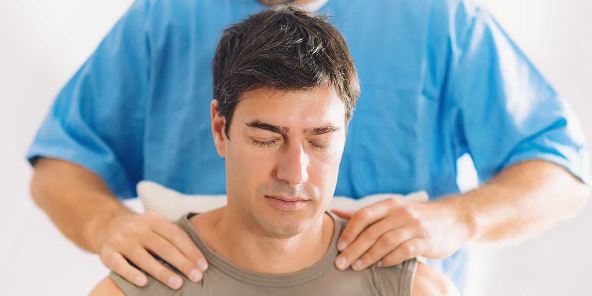A chiropractor places hands on a male patient's shoulders, illustrating professional care and adjustments to correct a neck hump.